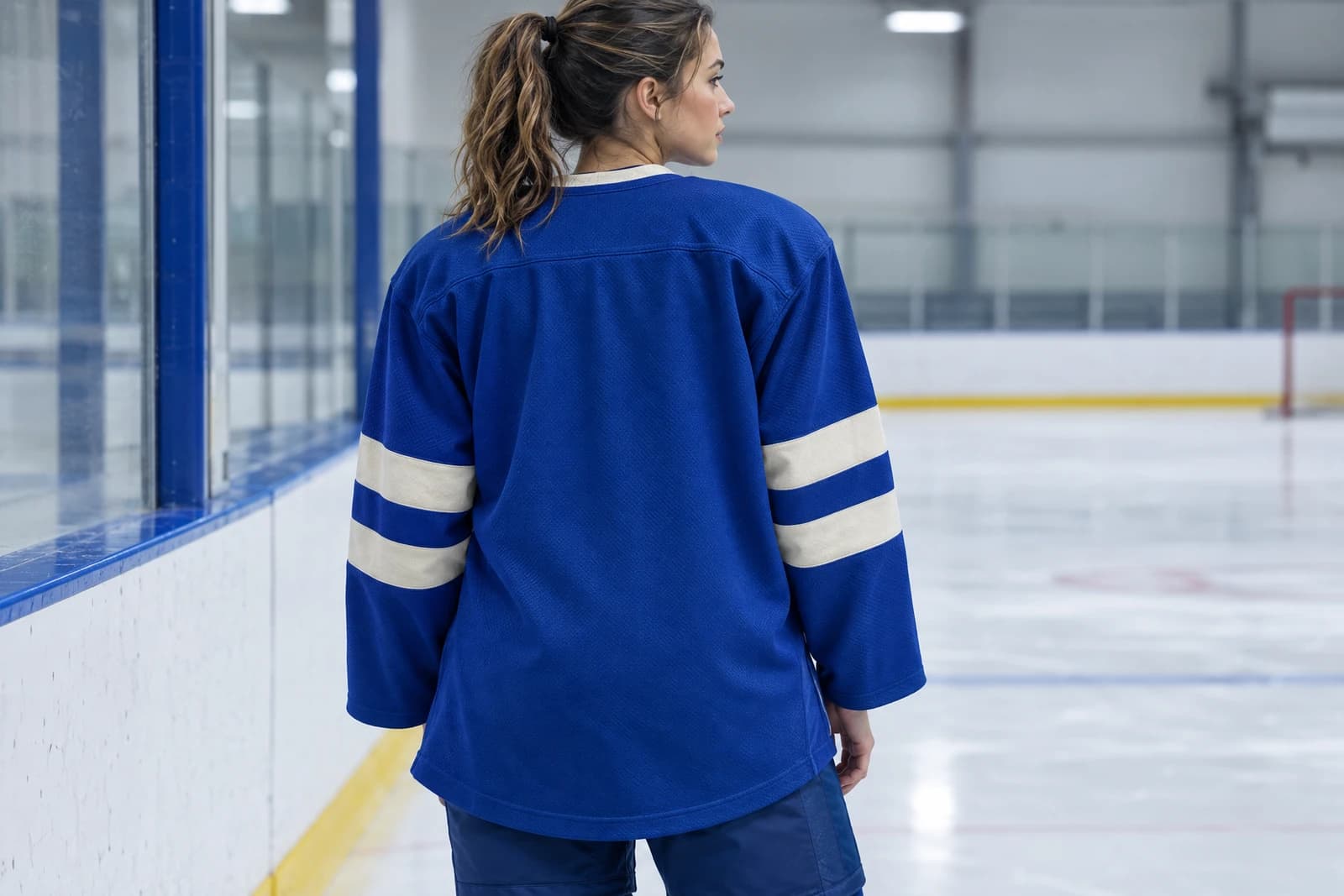 One adult seen from behind near the rink boards in a royal blue ice hockey jersey with cream sleeve stripes and a long hem