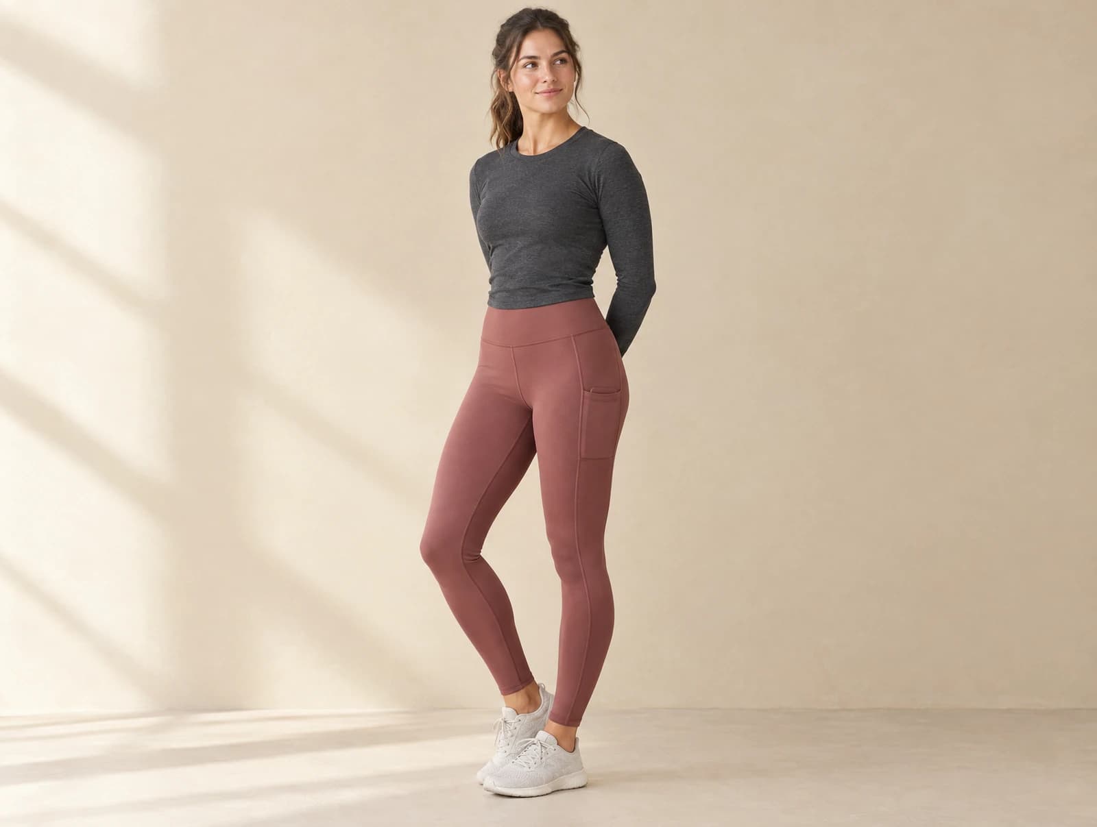 One adult standing in a bright studio wearing high-waisted mauve leggings with a visible thigh pocket and a fitted charcoal long-sleeve top