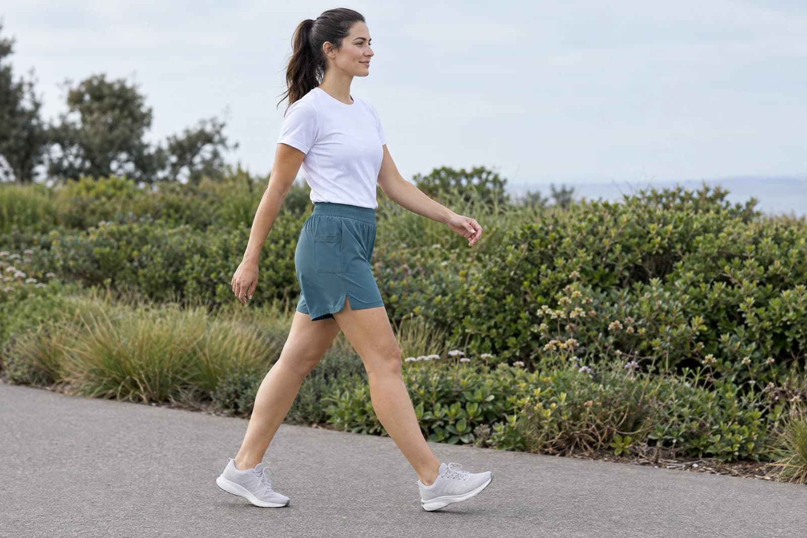 One adult walking along a paved coastal path in slate-teal athletic shorts with a gathered waistband and a side pocket