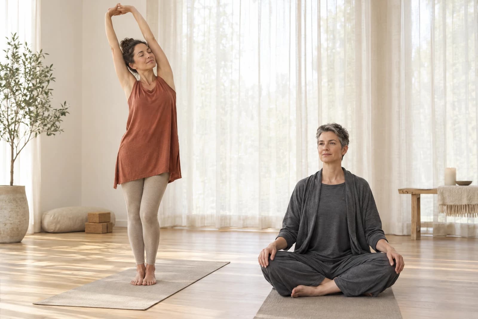 Two adults on yoga mats in a serene studio, one in a rust longline tank with oatmeal leggings and one seated in a grey knit lounge set